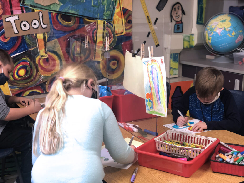 two students drawing with artwork clipped to plexiglass barriers