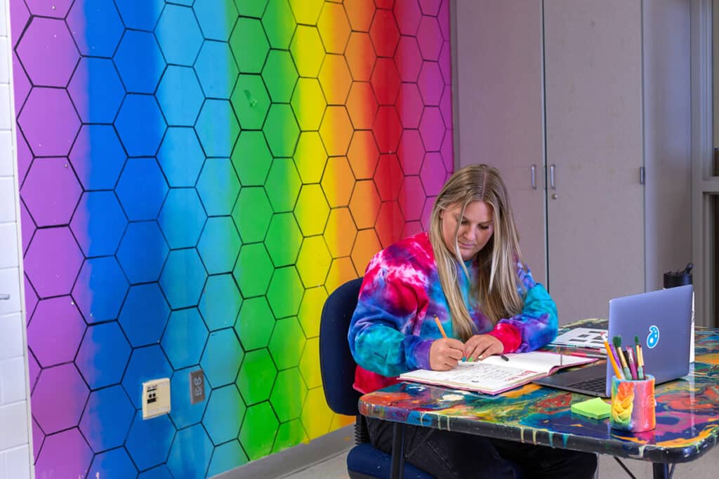 teacher in an art room writing in a sketchbook with a laptop